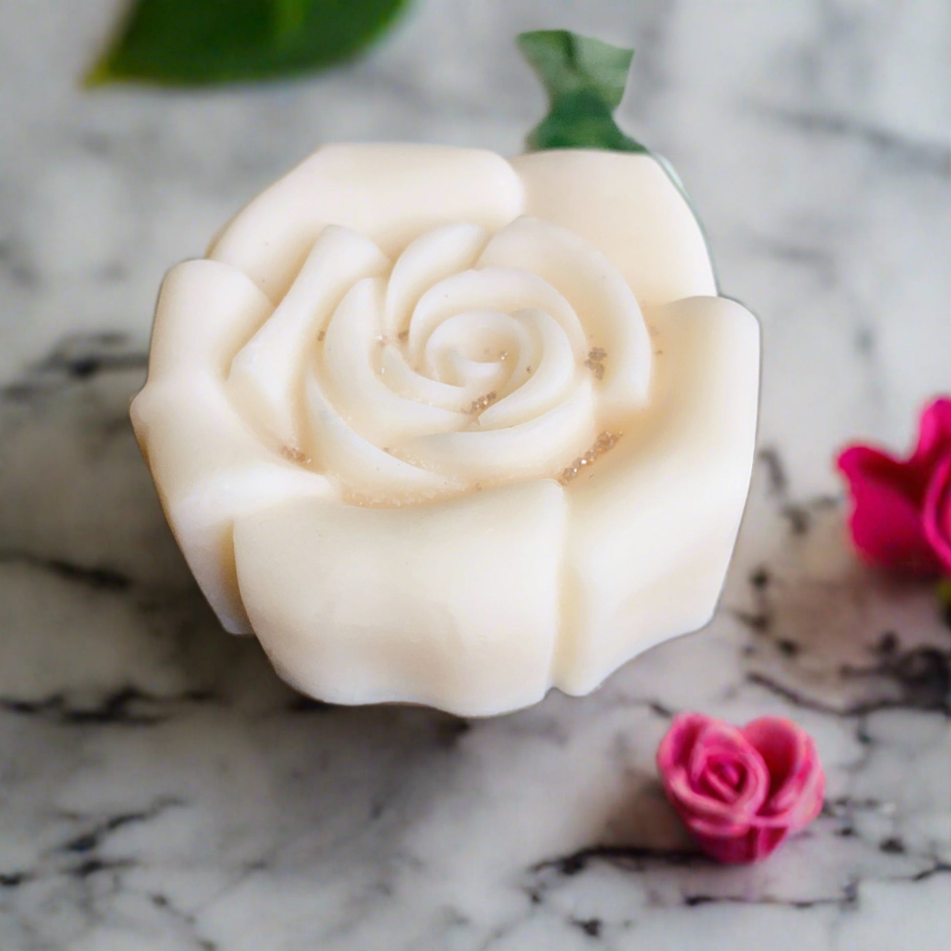 A white rose-shaped soap sits on marble with a green leaf and two pink Rose Candle Tart Melts from CeeCee Candle Company, adding elegance to the serene scene.