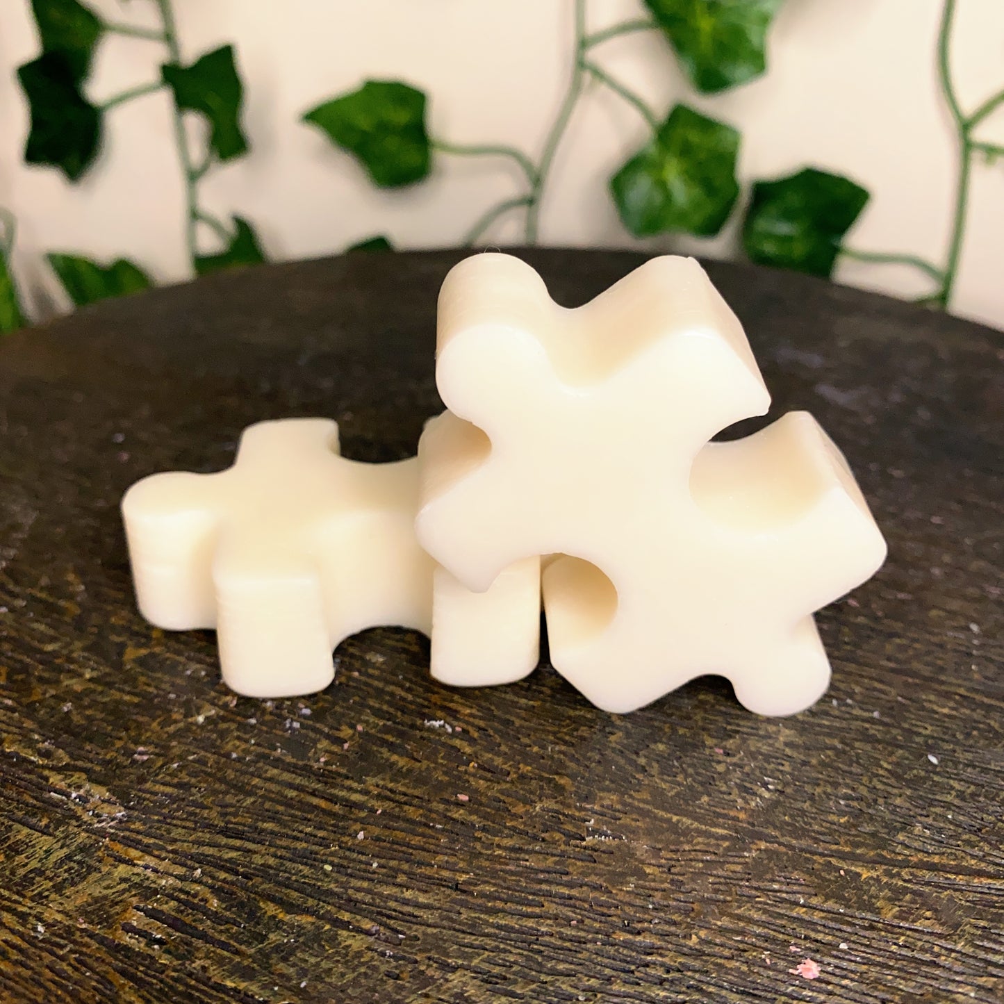Three creamy white Puzzle Pieces Wax Melts by CeeCee Candle Company rest on a textured wooden surface, creating a cozy atmosphere. Two are stacked, with blurred leafy vines in the background.