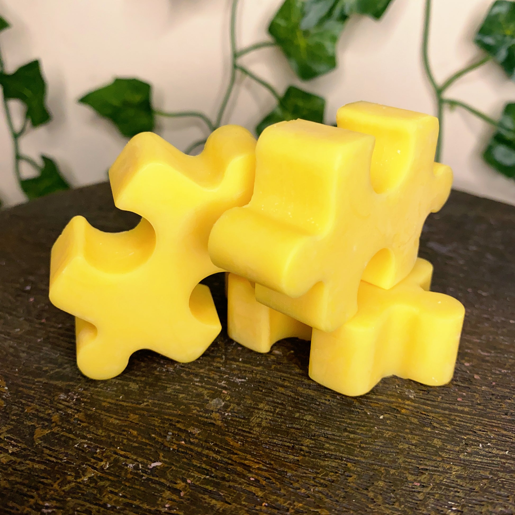 Three yellow Puzzle Pieces Wax Melts from CeeCee Candle Company are stacked on a dark wooden surface, with green leafy vines blurred in the background.